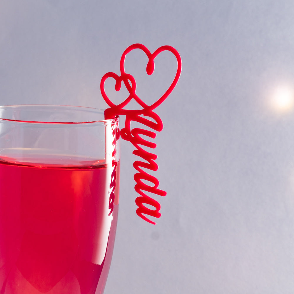 Gros plan sur un verre décoré d’un marque-verre Double Cœur rouge personnalisé “Lynda”, accroché au bord du verre rempli d’une boisson rose.