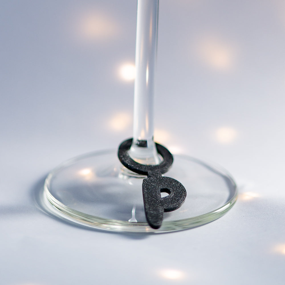 Marque-verre noir en forme de grande initiale glissée autour du pied d’un verre à pied, accessoire personnalisé pour identifier facilement les verres.