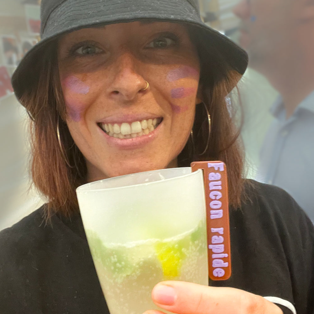 Personne souriante tenant un verre décoré d’un marque-verre Indien “Faucon rapide” lors d’une fête à thème.