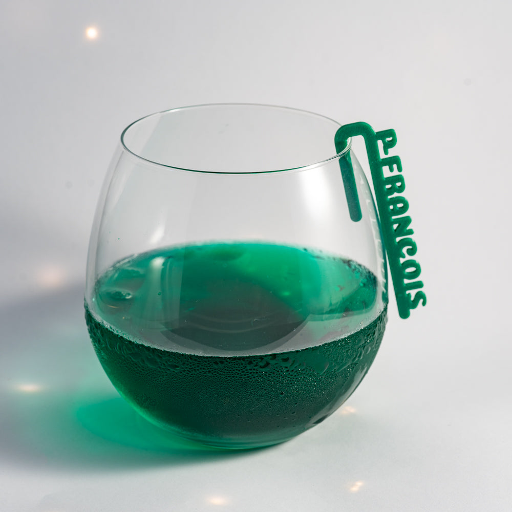 Marque-verre Linéal en PLA vert, formé d’une barre fine avec un prénom écrit en lettres verticales en relief. Le clip est fixé sur le bord d’un verre transparent de forme arrondie, rempli d’une boisson verte. La surface extérieure du verre présente de la condensation, donnant un aspect frais.