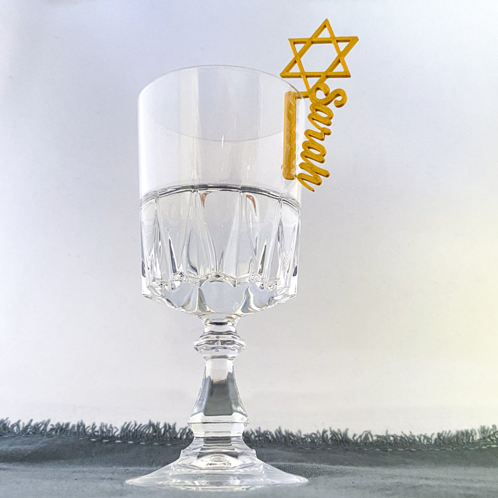 Verre à pied avec un marque-verre doré en forme d’Étoile de David et prénom “Sarah” accroché sur le bord.