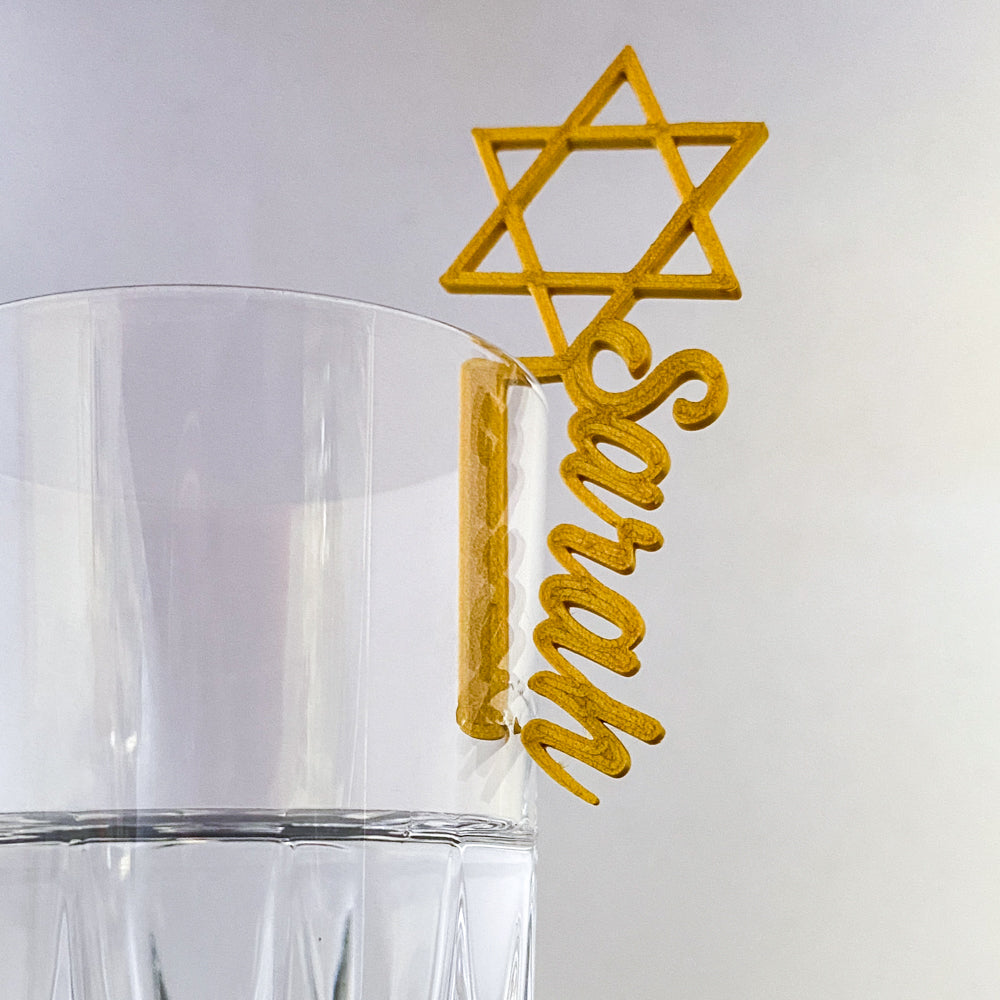 Marque-verre doré en forme d’Étoile de David avec le prénom “Sarah”, accroché au bord d’un verre transparent.