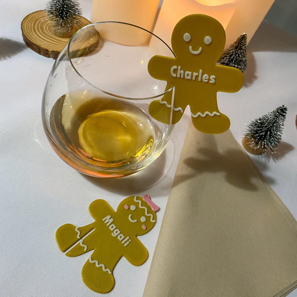 Verres posés sur une table décorée pour Noël, chacun accompagné d’un marque-verre Bonhomme de pain d'épices en PLA couleur miel portant les prénoms “Charles” et “Magali”. Les petits personnages souriants, décorés de détails blancs et roses, sont clipsés ou posés près des verres, entourés de mini sapins et de lumières chaleureuses.