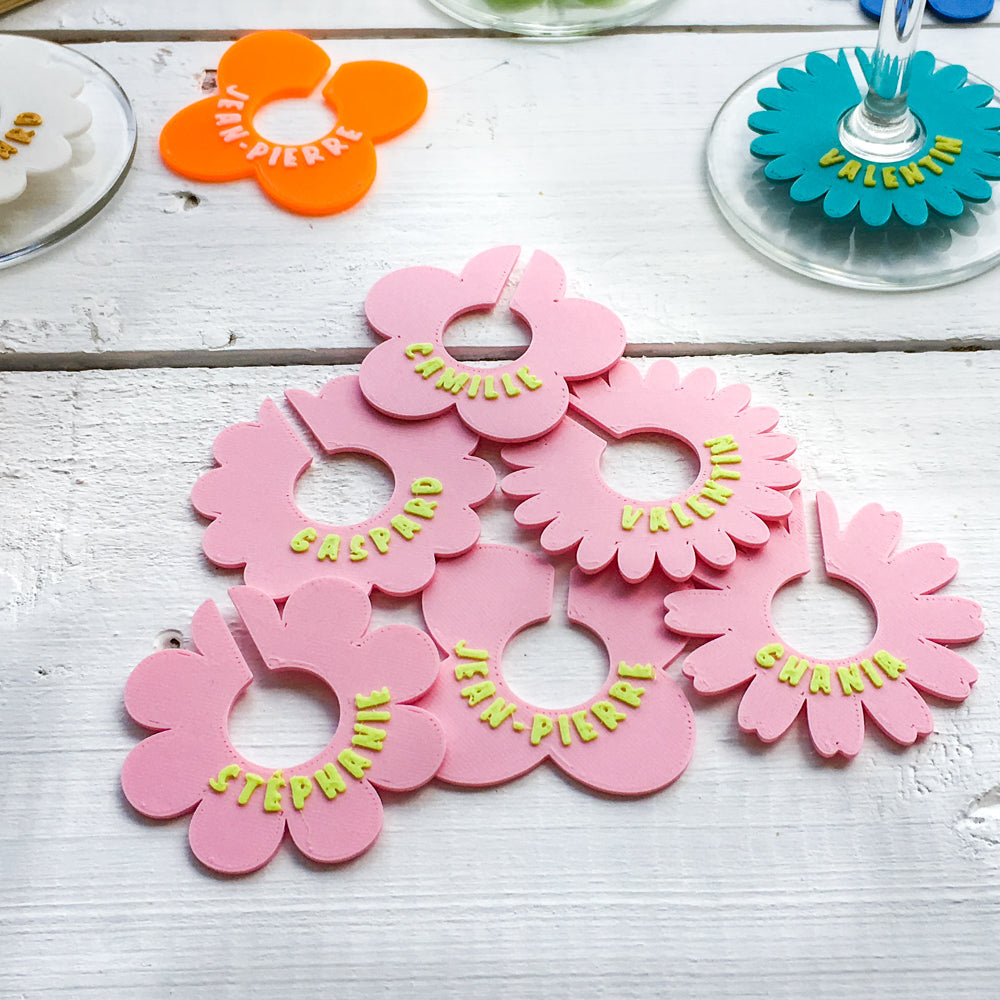 Marque-verres en forme de fleurs roses personnalisées avec des prénoms, disposés sur une table en bois blanche pour un apéritif coloré et convivial.