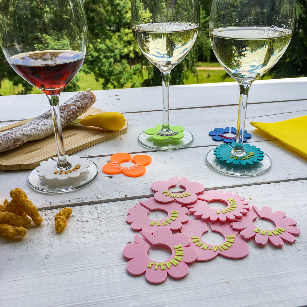 Marque-verres en forme de fleurs colorées personnalisées avec les prénoms, disposés autour de verres à pied lors d’un apéritif en extérieur.