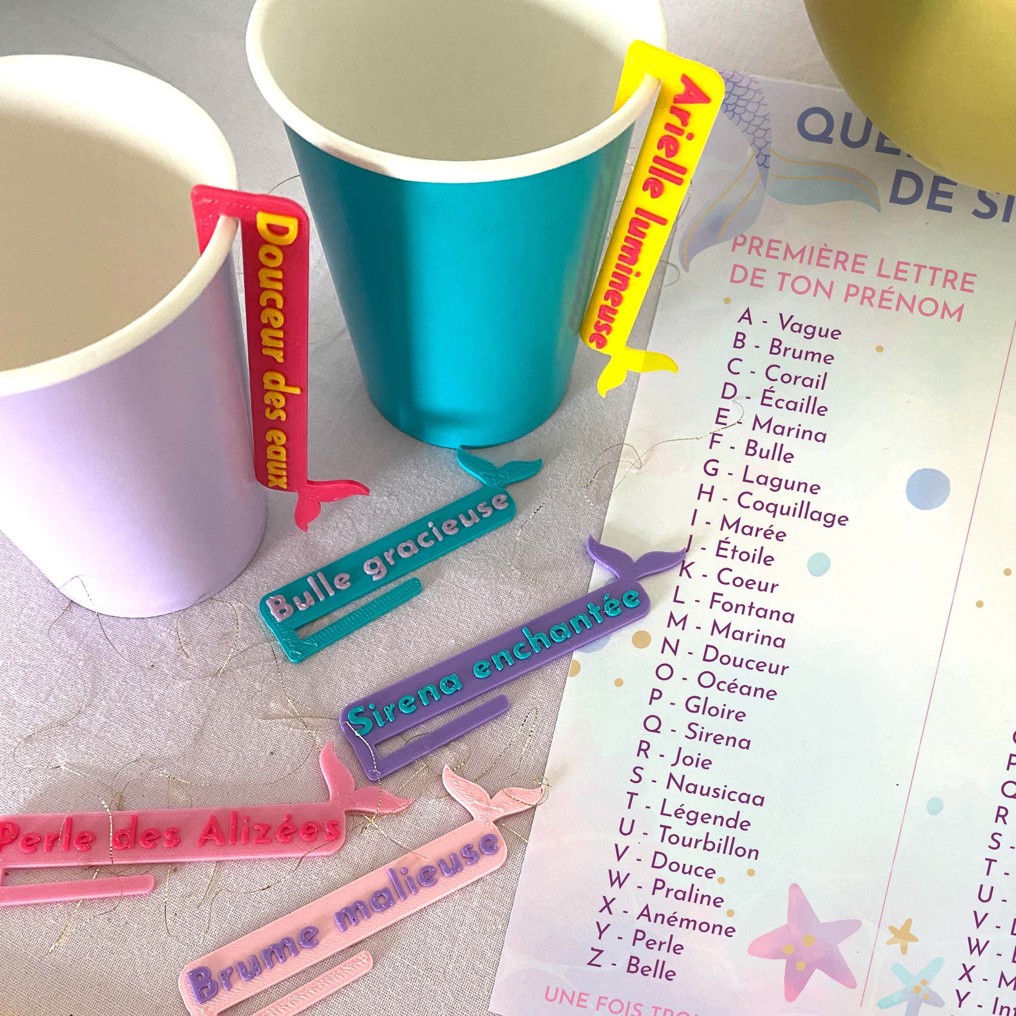 Marque-verres Sirènes colorés posés autour de gobelets, avec la feuille  “Quel est ton nom de Sirène ?” en arrière-plan et des étiquettes aux noms féeriques comme Arielle lumineuse, Douceur des eaux ou Brume malicieuse.