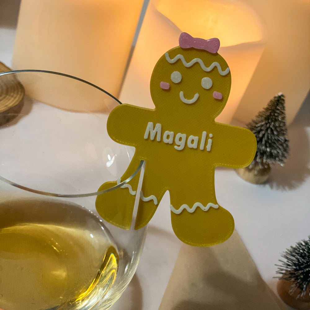 Gros plan sur un verre décoré d’un marque-verre Bonhomme de pain d’épices personnalisé “Magali”, posé devant des bougies lumineuses et de petites décorations hivernales, créant une ambiance chaleureuse et festive.