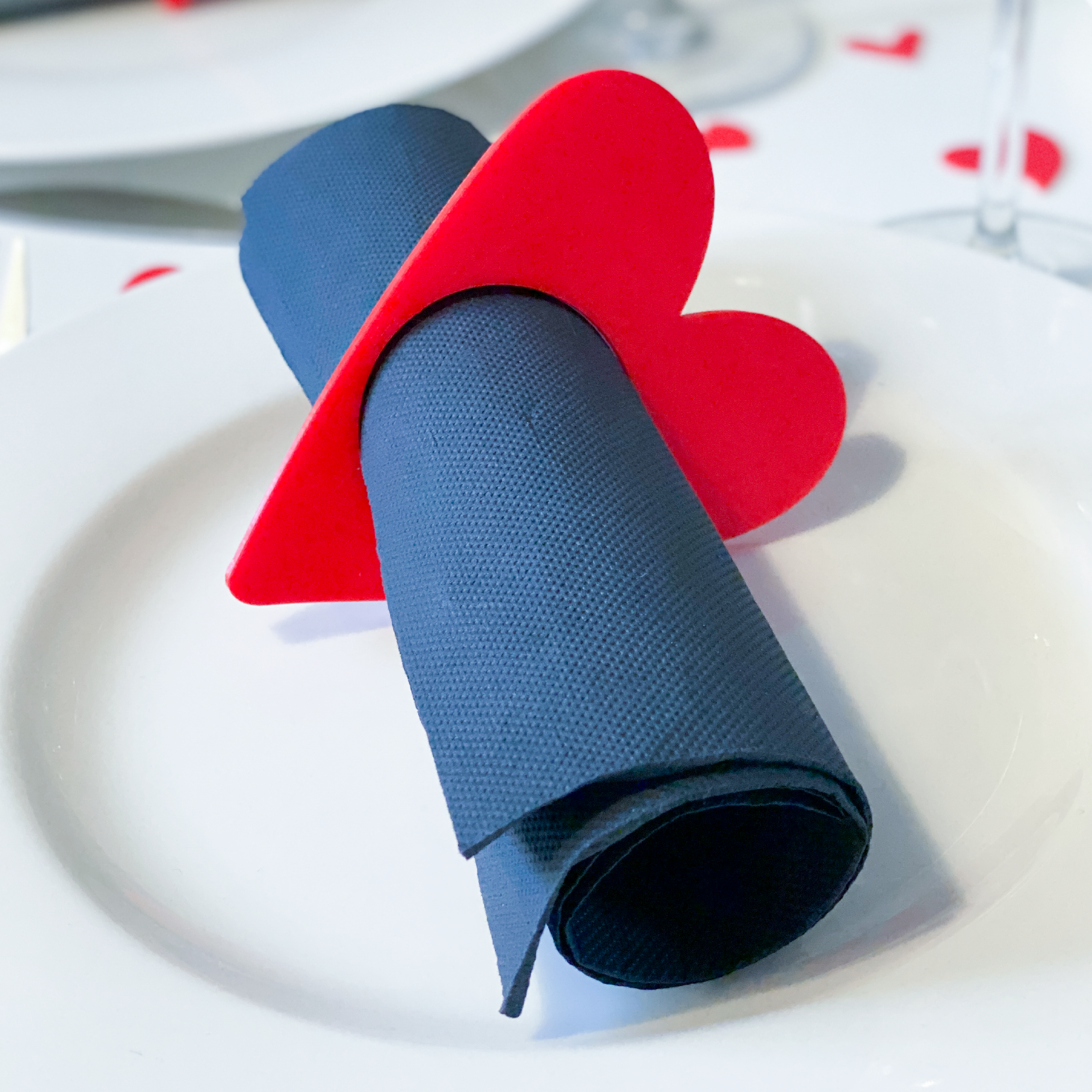 Rond de serviette en forme de cœur rouge tenant une serviette noire sur une assiette blanche, décor de table romantique.