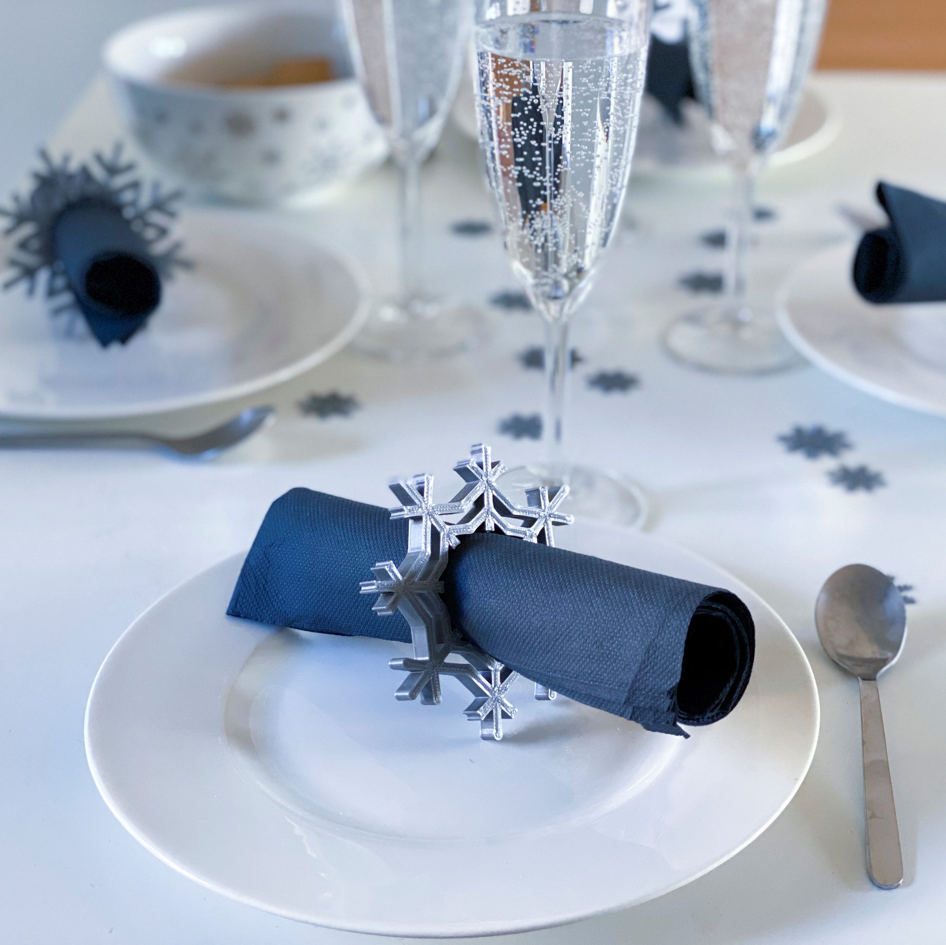 Rond de serviette en forme de flocon de neige argenté posé autour d’une serviette bleu nuit sur une assiette blanche, avec d’autres flocons décoratifs dispersés sur une table d’hiver élégante.