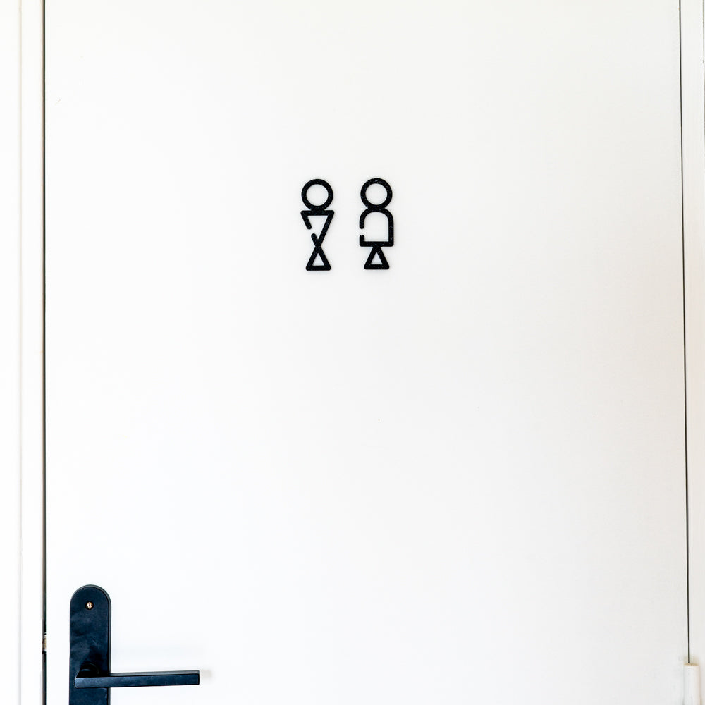 Signalétique toilettes stylisée noire collée sur une porte blanche, pictogrammes homme et femme géométriques au-dessus d’une poignée de porte noire.