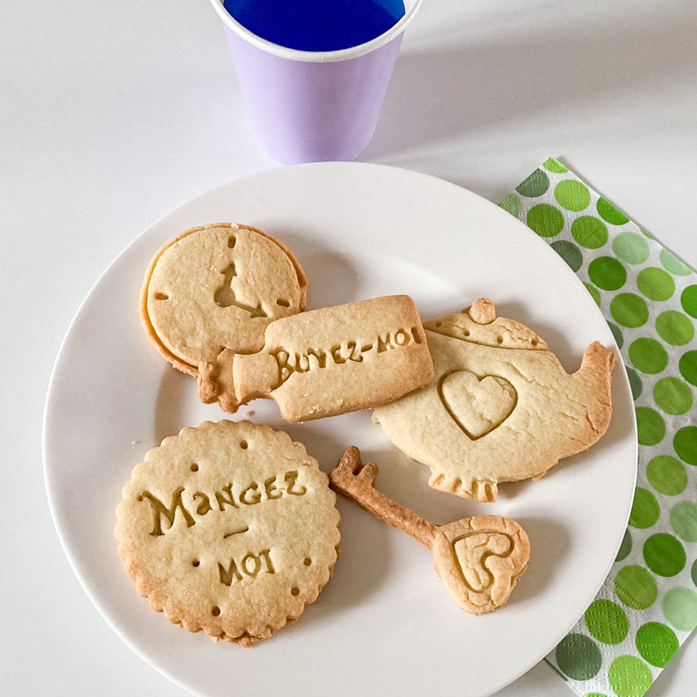 Assiette de biscuits maison inspirés d’Alice au pays des merveilles, en forme de montre, bouteille "Buvez-moi", clé et théière, réalisés avec des emporte-pièces violets.