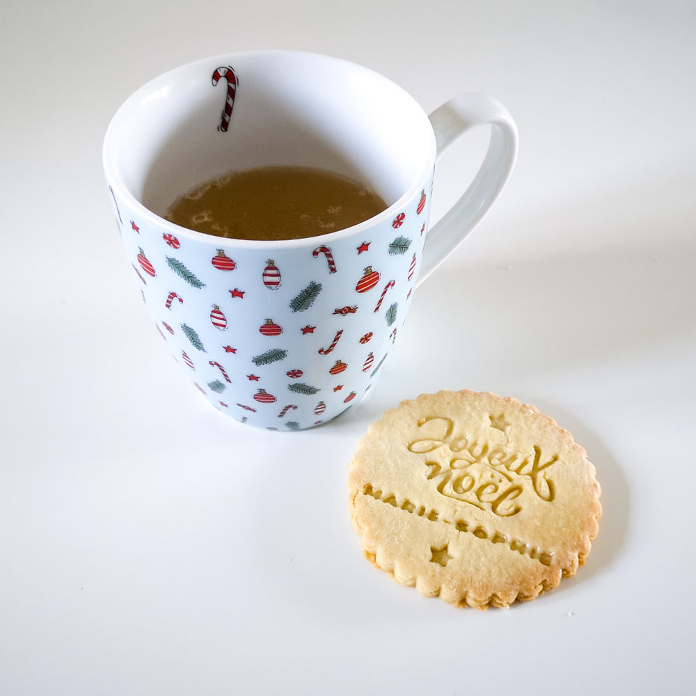 Biscuit rond Joyeux Noël personnalisé avec prénom et tasse décorée de motifs de fête sur fond blanc.