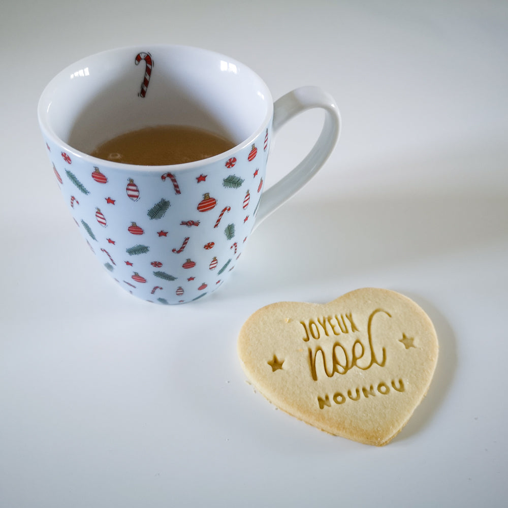 Biscuit en forme de cœur marqué Joyeux Noël avec prénom personnalisé, réalisé avec un emporte-pièce sur pâte à biscuits, à côté d'une tasse aux motifs de Noël.