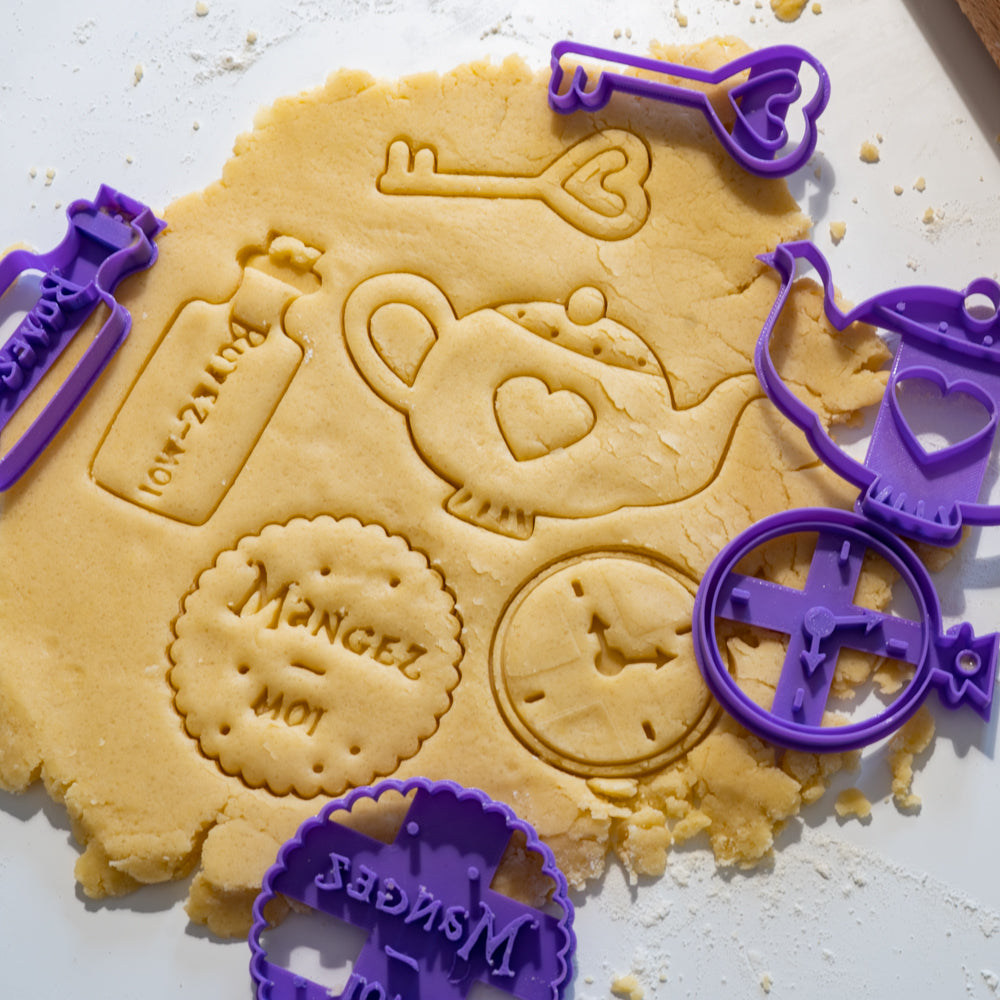 Lot d’emporte-pièces Alice au pays des merveilles imprimant une clé, une théière, une horloge, un biscuit “Mangez-moi” et une fiole “Buvez-moi” dans une pâte à biscuits.