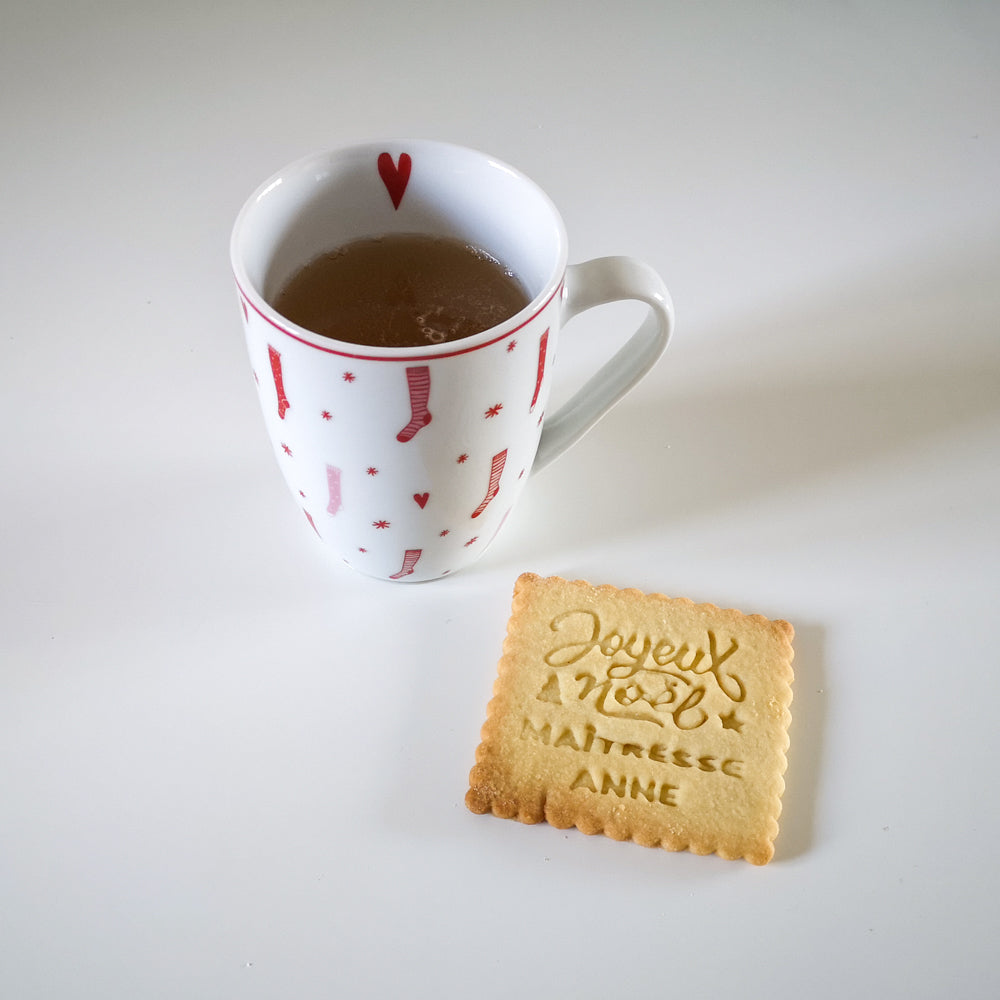 Biscuit Joyeux Noël personnalisé pour maîtresse posé à côté d'une tasse de thé sur une table blanche.
