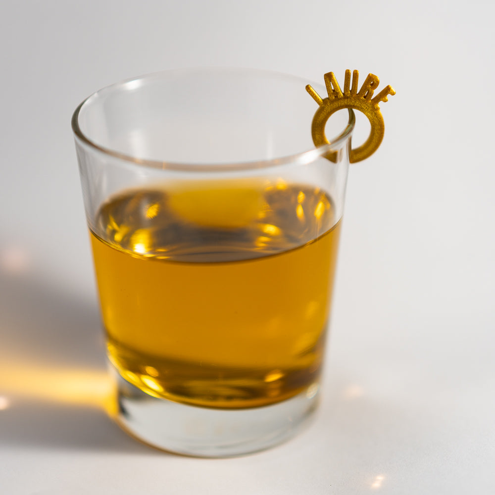 Marque-verre en forme d’anneau doré, portant le prénom “Laure” en lettres verticales, accroché au bord d’un verre transparent rempli d’une boisson ambrée.