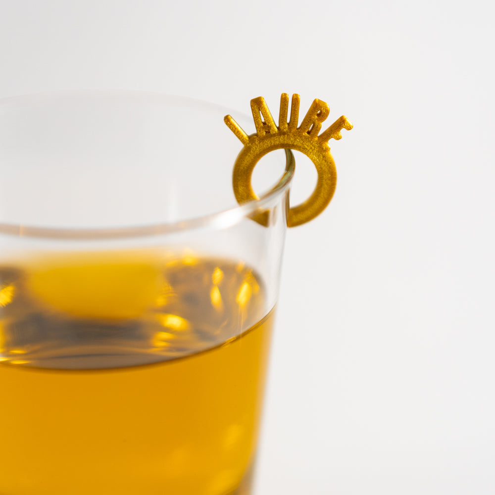 Marque-verre doré en forme d’anneau doré avec les lettres du prénom “Laure” dressées autour du cercle, clipsé sur le bord d’un verre rempli d’une boisson ambrée.