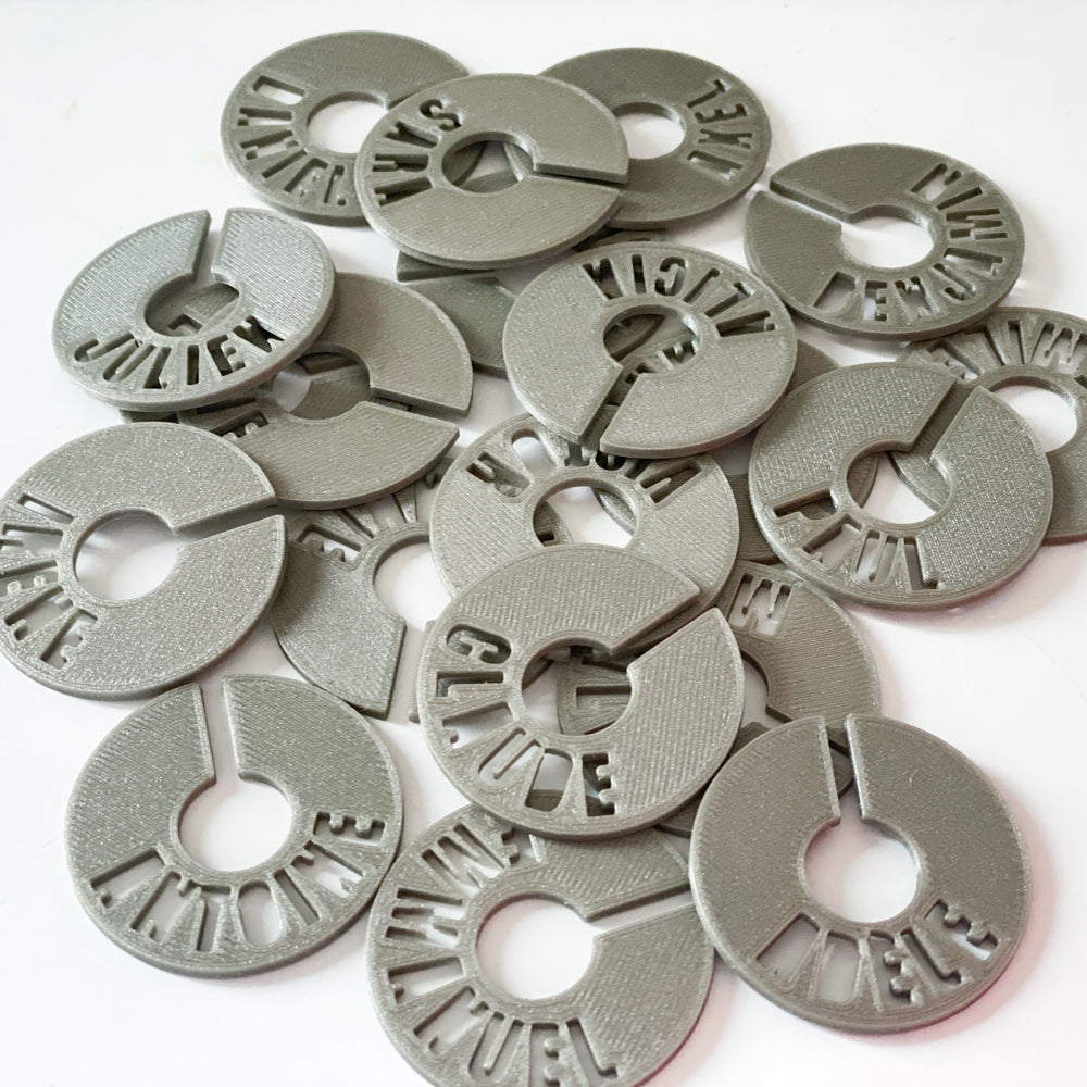 Ensemble de marque-verres creusés en PLA gris argent, empilés en vrac, chacun avec un prénom découpé en arc de cercle autour de l’ouverture permettant de les glisser autour du pied du verre.