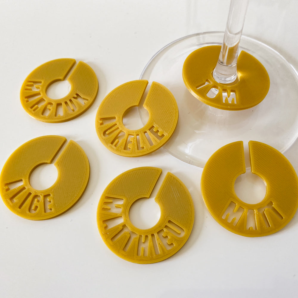 Ensemble de marque-verres creusés en PLA  couleurs Miel, en forme de rond fendu, posés autour de verres à pied. Chaque pastille affiche un prénom découpé dans la matière : Margaux, Aurélie, Tom, Alice, Mathieu et Matt.