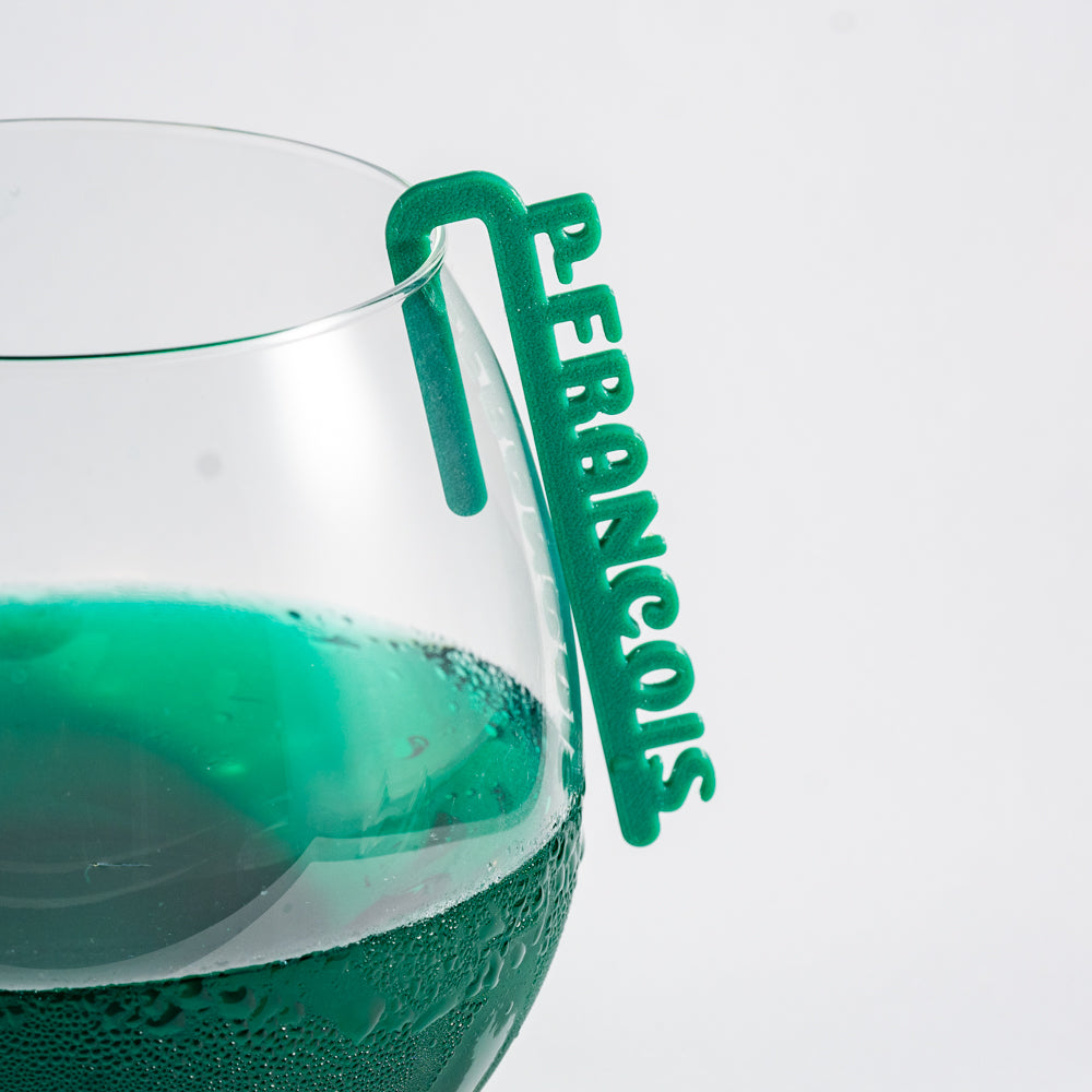 Marque-verre Linéal en plastique PLA vert, formé d’une barre fine avec des lettres verticales en relief indiquant un prénom. Le clip est fixé sur le bord d’un verre transparent rempli d'une boisson verte. Les lettres sont nettes et allongées, donnant un aspect minimaliste et moderne.