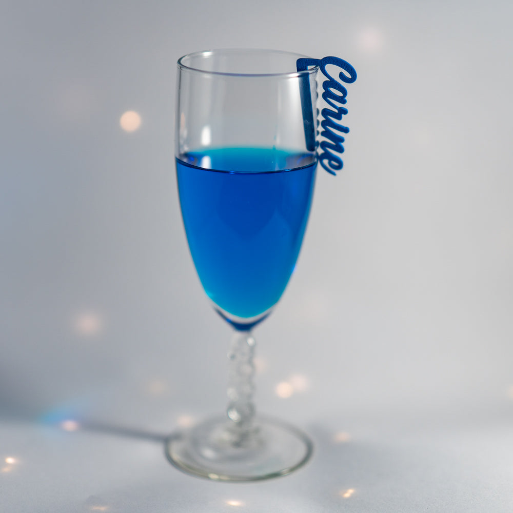 Flûte à champagne remplie d’une boisson bleue vive, posée sur un fond clair légèrement scintillant. Un marque-verre Originel bleu, en écriture cursive et portant le prénom “Carine”, est clipsé sur le bord du verre. La mise en scène est lumineuse et épurée, avec quelques reflets doux autour du pied de la flûte.