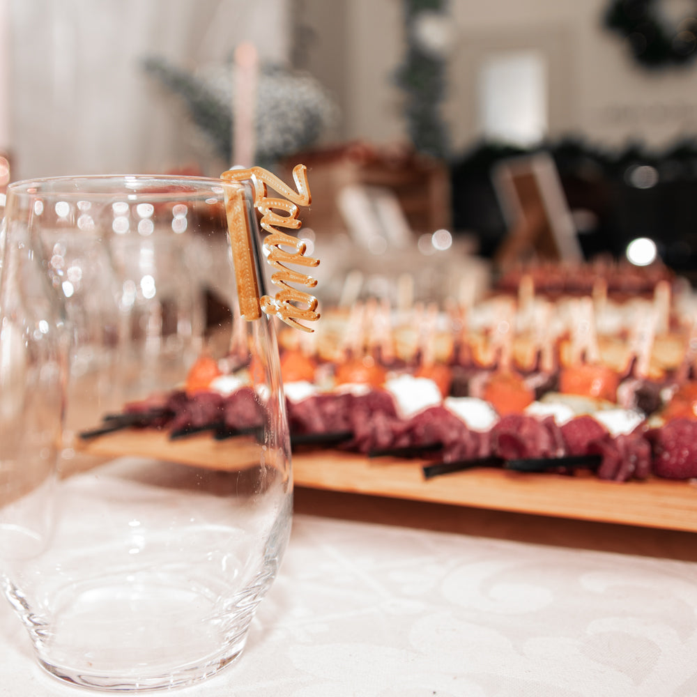 Verre transparent posé sur une table de réception, avec un marque-verre Originel doré portant le prénom “Zadig” clipsé sur son bord. En arrière-plan, légèrement flou, un buffet coloré de brochettes apéritives crée une ambiance chaleureuse et festive.
