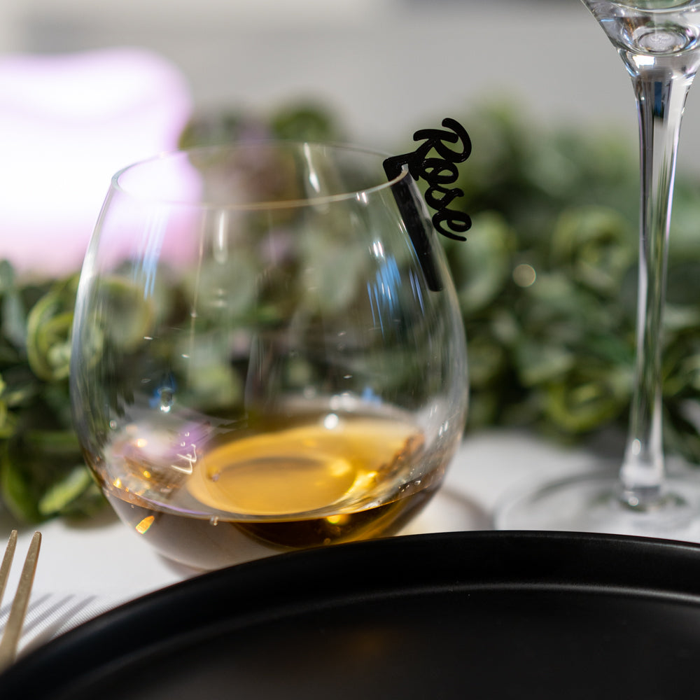 Verre rond type whisky rempli d’une boisson ambrée, posé sur une table élégante. Un marque-verre Originel noir, en écriture cursive, portant le prénom “Rose”, est clipsé au bord du verre. En arrière-plan, une décoration végétale floutée crée une ambiance chic et douce.