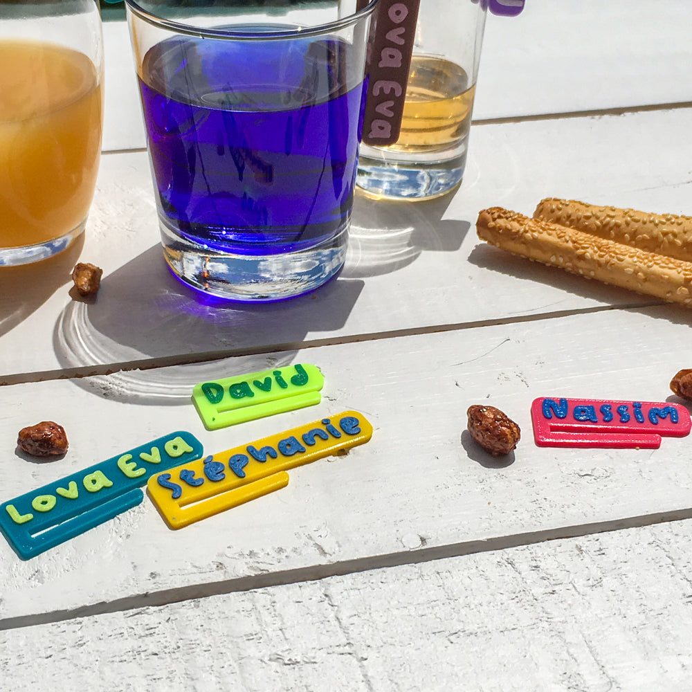 Gros plan sur des marque-verres bicolores posés sur une table en bois clair, avec les prénoms “Lova Eva”, “Stéphanie”, “David” et “Nassim” en lettres en relief colorées. Boissons et gressins visibles en arrière-plan, ambiance apéritive en extérieur.