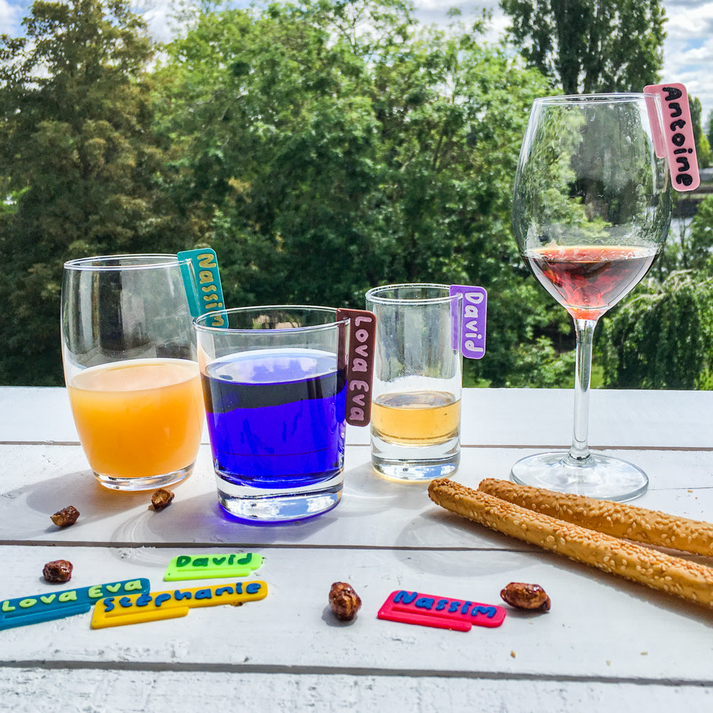 Verres posés en extérieur avec des marque-verres colorés clipsés au bord : prénoms en relief sur des  marque-verres bicolores. Plusieurs boissons colorées (orange, bleu, rosé) et marque-verres supplémentaires posés sur la table. Ambiance apéritive en plein air, arbres en arrière-plan.