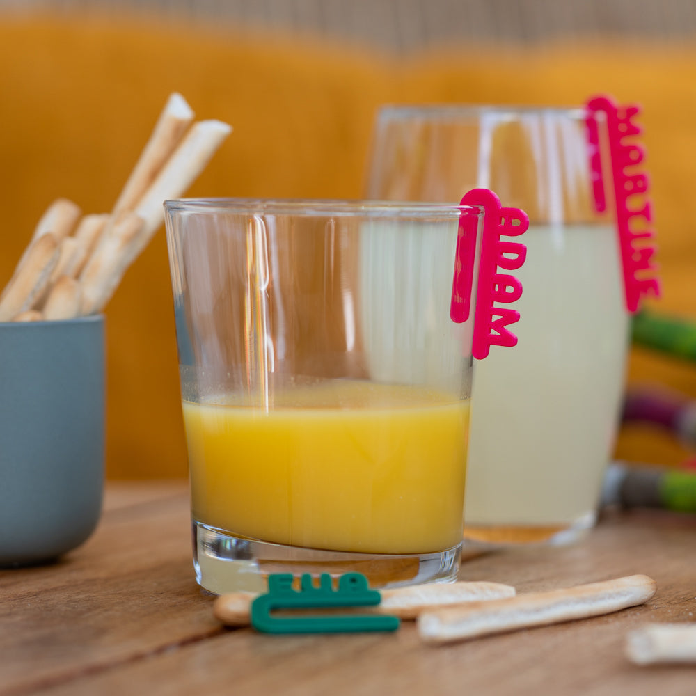 Deux verres de jus posés sur une table, chacun équipé d’un marque-verre Linéal rose portant les prénoms “Adam” et “Martine”. Au premier plan, un clip vert repose sur la table. L’ensemble crée une ambiance conviviale et colorée.