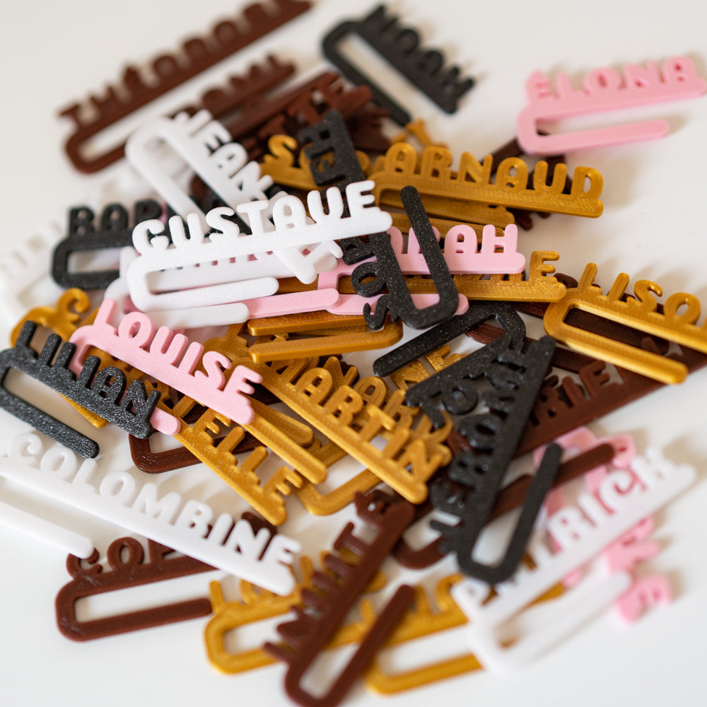 Pile de clips marque-verres Linéal personnalisés, avec prénoms écrits en lettres droites alignées sur une barre horizontale. Les pièces sont imprimées en PLA dans plusieurs coloris — blanc, noir, brun, or, rose — et entassées les unes sur les autres sur un fond clair.